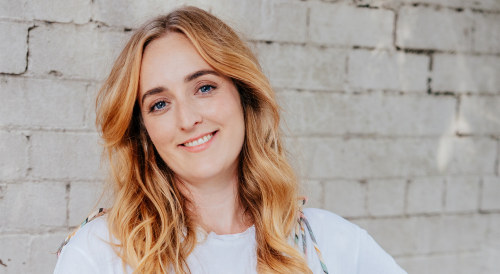 An author event phot of Amy Lovat. She is smiling at the camera in front of a white brick wall.